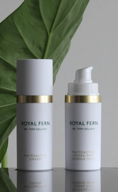 Produktfotografie von zwei Kosmetikprodukten der Marke Royal Fern, die vor einem großen grünen Blatt stehen. Links eine Creme mit Deckel, rechts eine Maske mit Pumpspender.