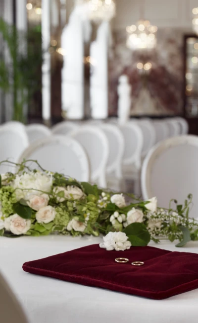 Zwei goldene Eheringe liegen auf einem bordeauxroten Kissen neben einem weißen Blumenarrangement in einem festlich dekorierten Saal.