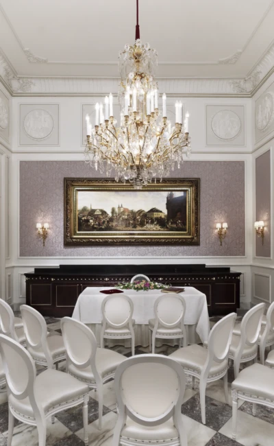 Hochzeitlich, gedeckter Raum mit weißen Stühlen, Kristalllüster und großem Gemälde an der Wand.