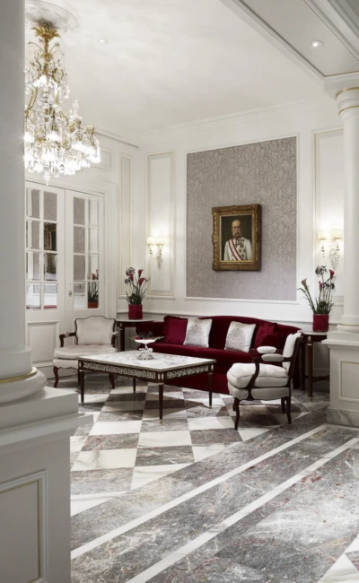 Eleganter Salon mit rotem Sofa, Marmorboden, Kristalllüster und Porträt an der Wand.