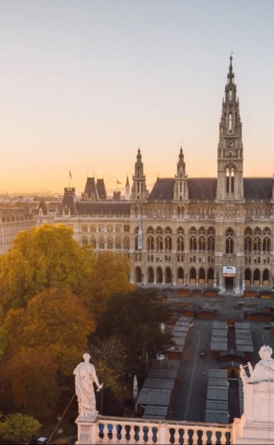 Wiener Rathaus im warmen Licht der untergehenden Sonne. Die neugotische Fassade des Gebäudes ist von zahlreichen Türmen und Verzierungen geprägt, wobei der mittlere Uhrturm besonders hervorsticht. Davor sind zahlreiche Marktstände aufgebaut – vermutlich für einen Markt oder ein bevorstehendes Fest. Im Vordergrund rahmen zwei weiße Statuen auf einem Balkon die Szene ein.