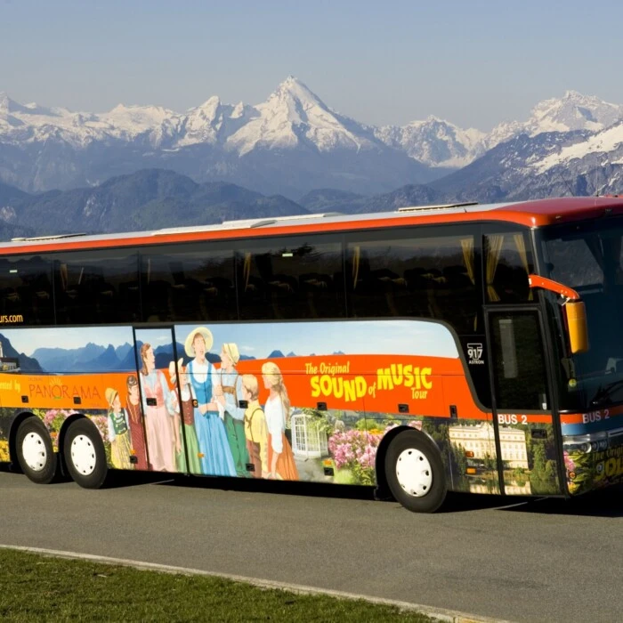 Ein bunter Reisebus der „Original Sound of Music Tour“ vor eindrucksvoller Bergkulisse mit schneebedeckten Gipfeln. Der Bus zeigt Illustrationen aus dem Film „The Sound of Music“ und steht auf einer Straße mit grüner Wiese im Vordergrund.