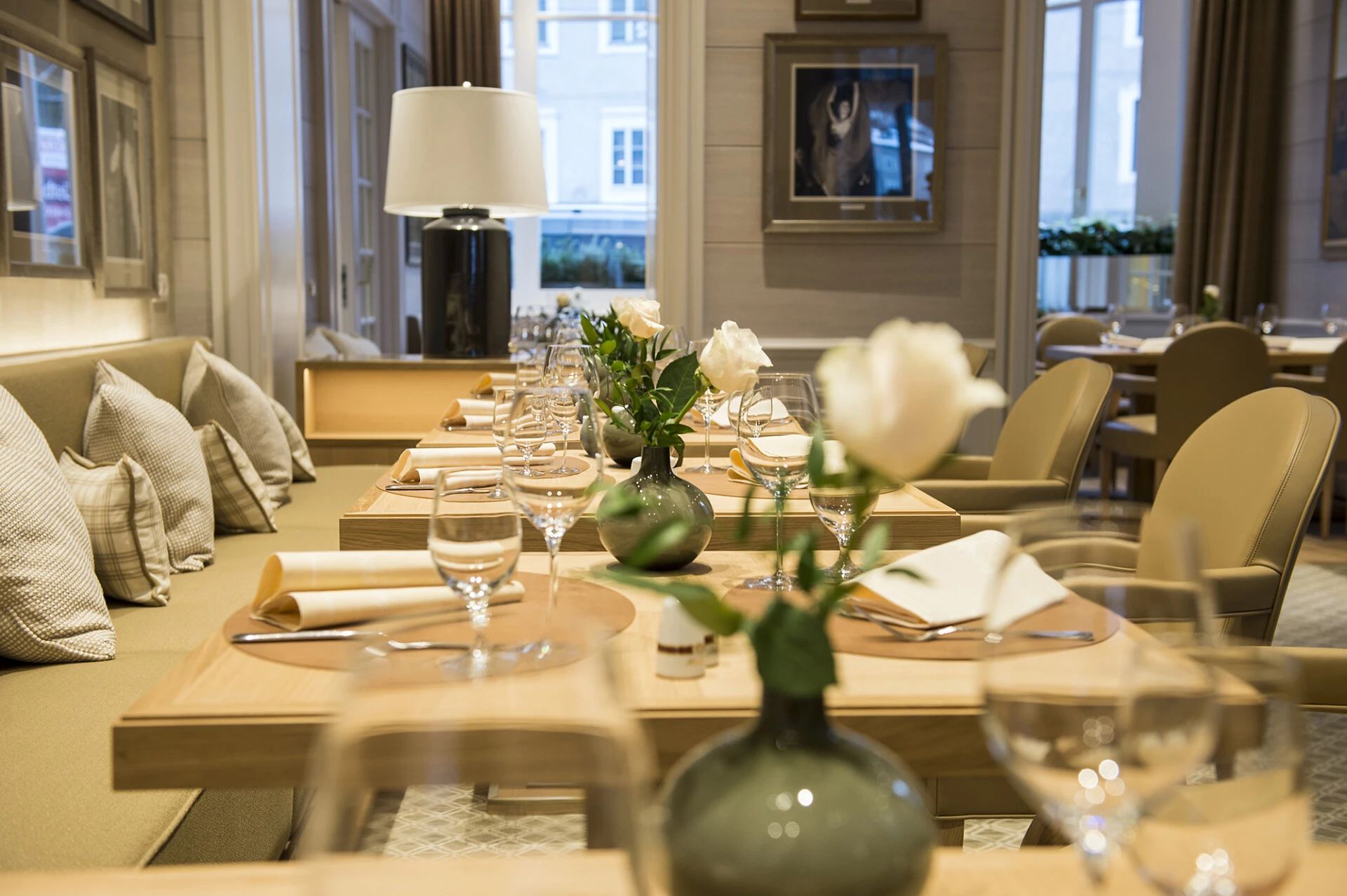 Elegant gedeckter Restaurantbereich mit hellen Holztischen, beigen Polstermöbeln und weißen Rosen in kleinen Vasen. Die Atmosphäre ist modern und stilvoll mit gemütlichem Flair.