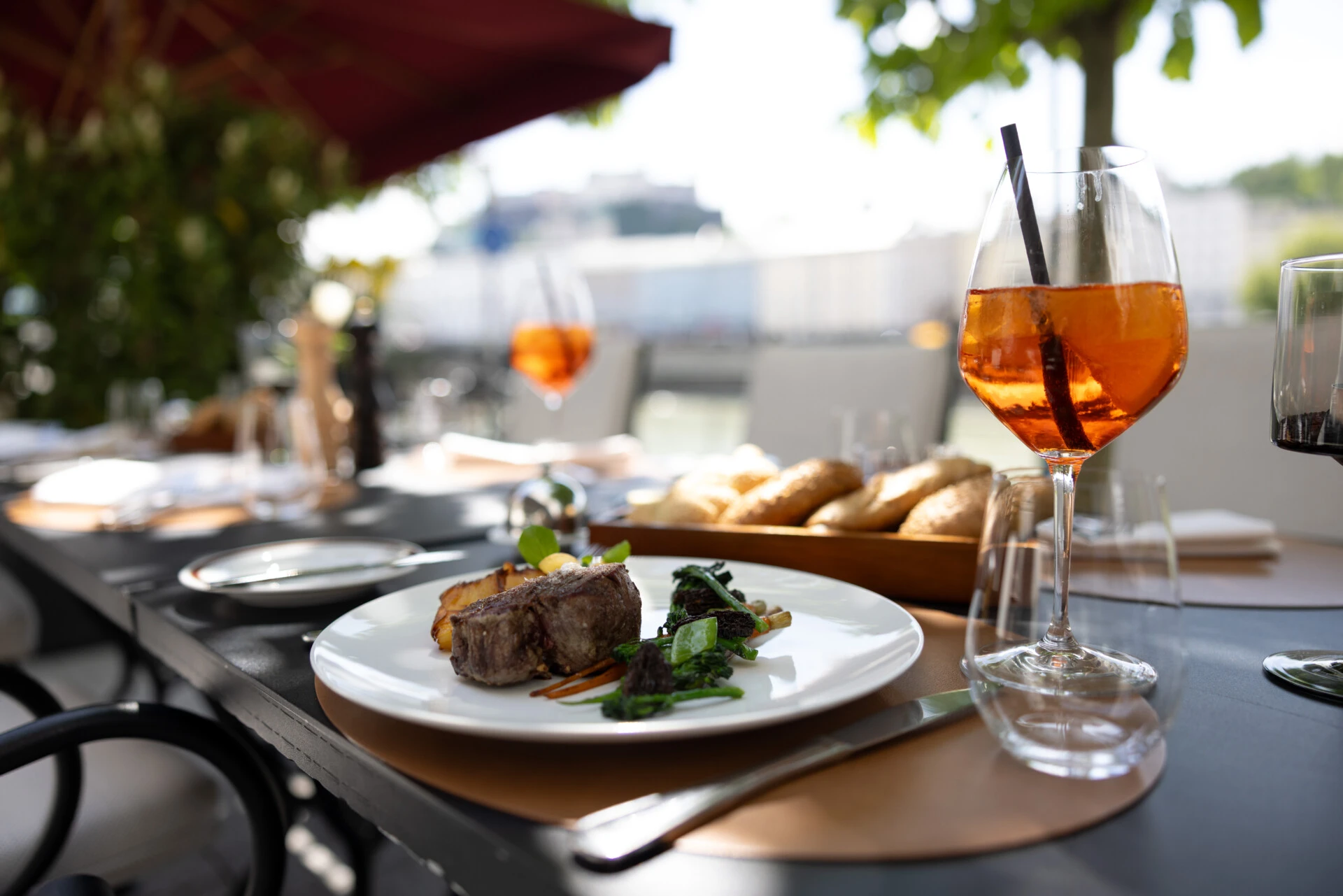 Ein elegant angerichtetes Mittagessen im Freien: zartes Rinderfilet mit Gemüse, knusprigen Kartoffeln und Brotvariation. Dazu ein Glas Aperitif – serviert im schattigen Gartenambiente. Im Hintergrund ist die Festung Hohensalzburg zu sehen.