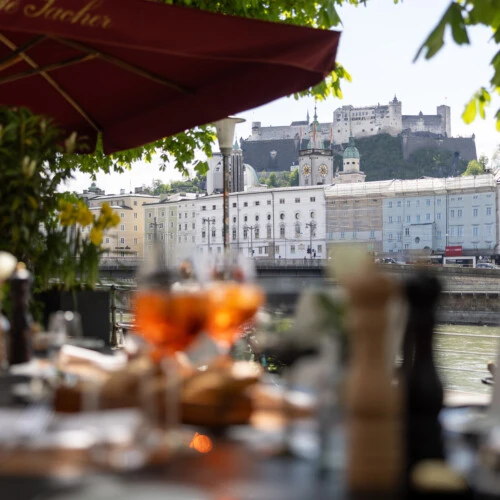 Terrasse des Café Sacher mit Blick auf die Salzburger Altstadt und die Festung Hohensalzburg – stilvoll gedeckte Tische, Aperitifs im Vordergrund, schattige Bäume und sommerliche Atmosphäre.