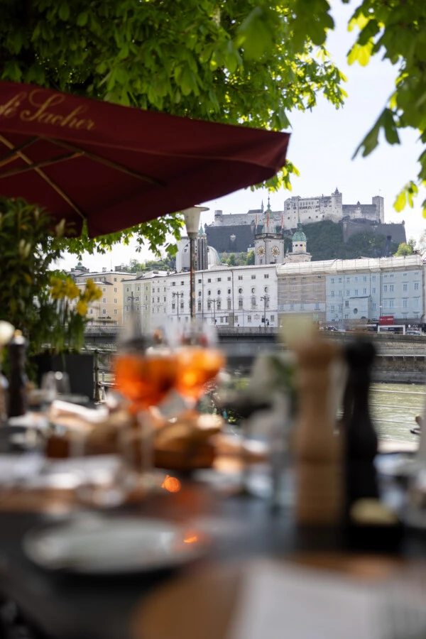 Terrasse des Café Sacher mit Blick auf die Salzburger Altstadt und die Festung Hohensalzburg – stilvoll gedeckte Tische, Aperitifs im Vordergrund, schattige Bäume und sommerliche Atmosphäre.