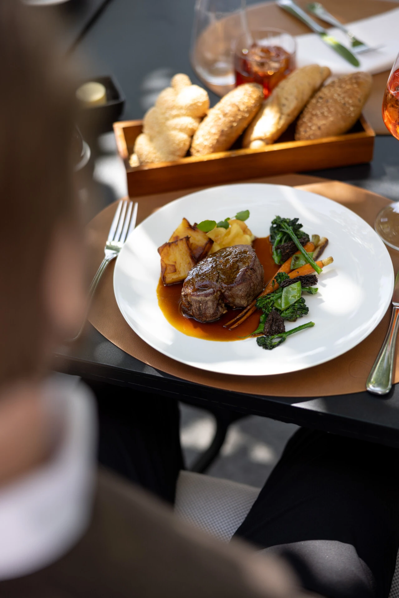 Ein elegant angerichtetes Mittagessen im Freien: zartes Rinderfilet mit Gemüse, knusprigen Kartoffeln und Brotvariation. Dazu ein Glas Aperitif – serviert im schattigen Gartenambiente.