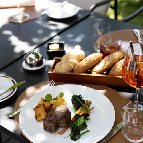 Ein elegant angerichtetes Mittagessen im Freien: zartes Rinderfilet mit Gemüse, knusprigen Kartoffeln und Brotvariation. Dazu ein Glas Aperitif – serviert im schattigen Gartenambiente.