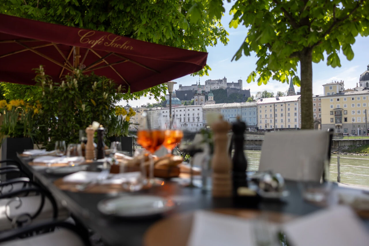 Ein stilvoll gedeckter Tisch im Café Sacher Salzburg unter einem roten Sonnenschirm mit Blick auf die Salzach und die Festung Hohensalzburg.
