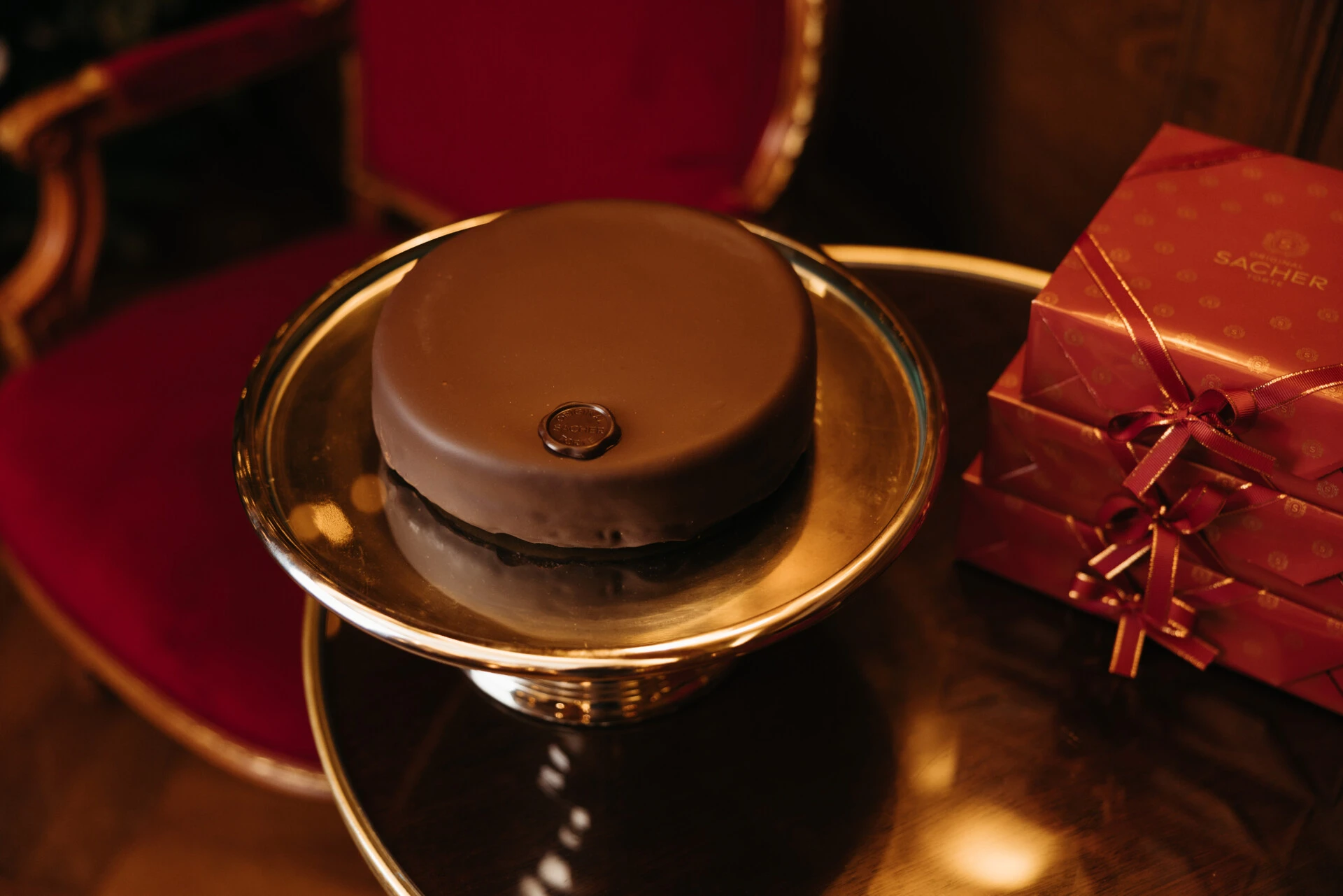 Die Original Sacher-Torte auf silberner Etagere, daneben edel verpackte Geschenkboxen mit rotem Schleifenband.