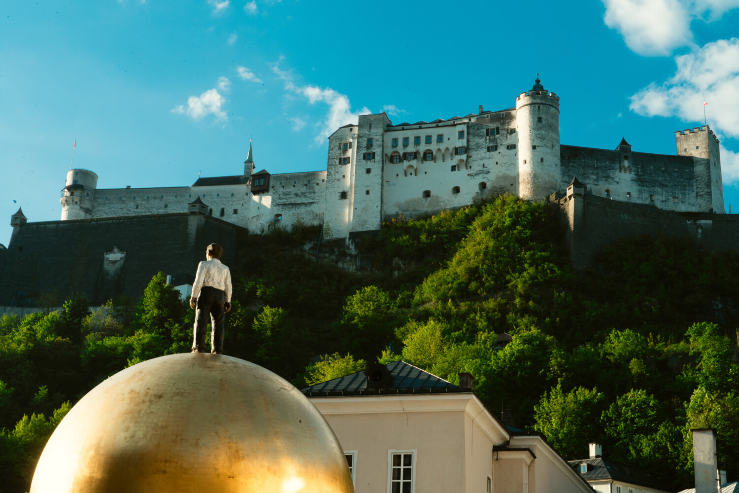 Die majestätische Festung Hohensalzburg, umgeben von üppigen Bäumen, thront stolz. In ihrer Nähe befindet sich die faszinierende Statue eines Kindes auf einer riesigen goldenen Kugel.
