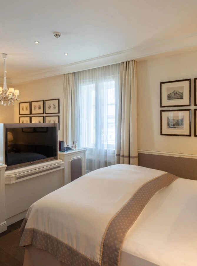 family-suite-hote-sacher-salzburg-bed-and-TV Geräumige Hotelsuite im Hotel Sacher mit Kingsize-Bett, gemütlicher Sitzecke und TV-Konsole vor dem Bett