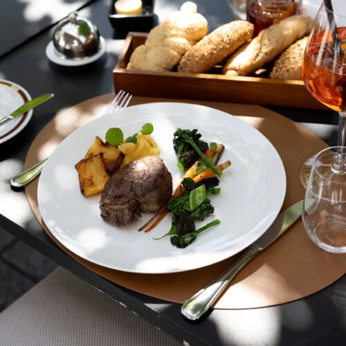 Feinstes Rinderfilet mit glasiertem Gemüse, Kartoffelgratin und Apfelpüree, serviert im eleganten Außenbereich mit Brotkorb und Aperitif.