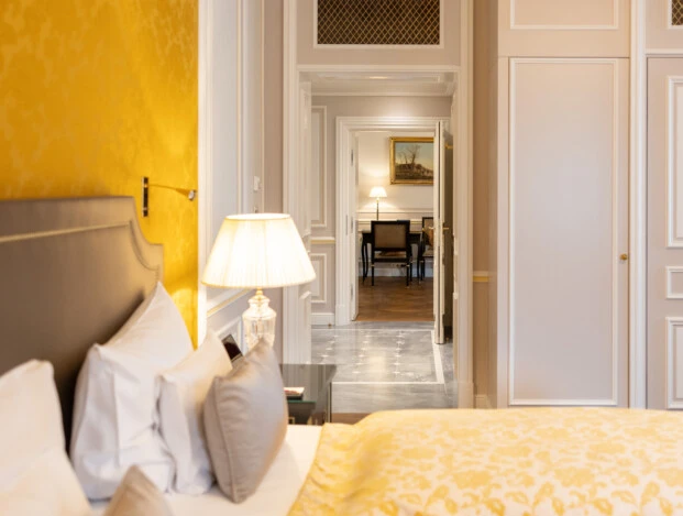 Blick vom Bett mit goldgelber Decke durch einen hellen Flur auf einen Schreibtischbereich, flankiert von klassischer Wandgestaltung und stilvoller Beleuchtung.