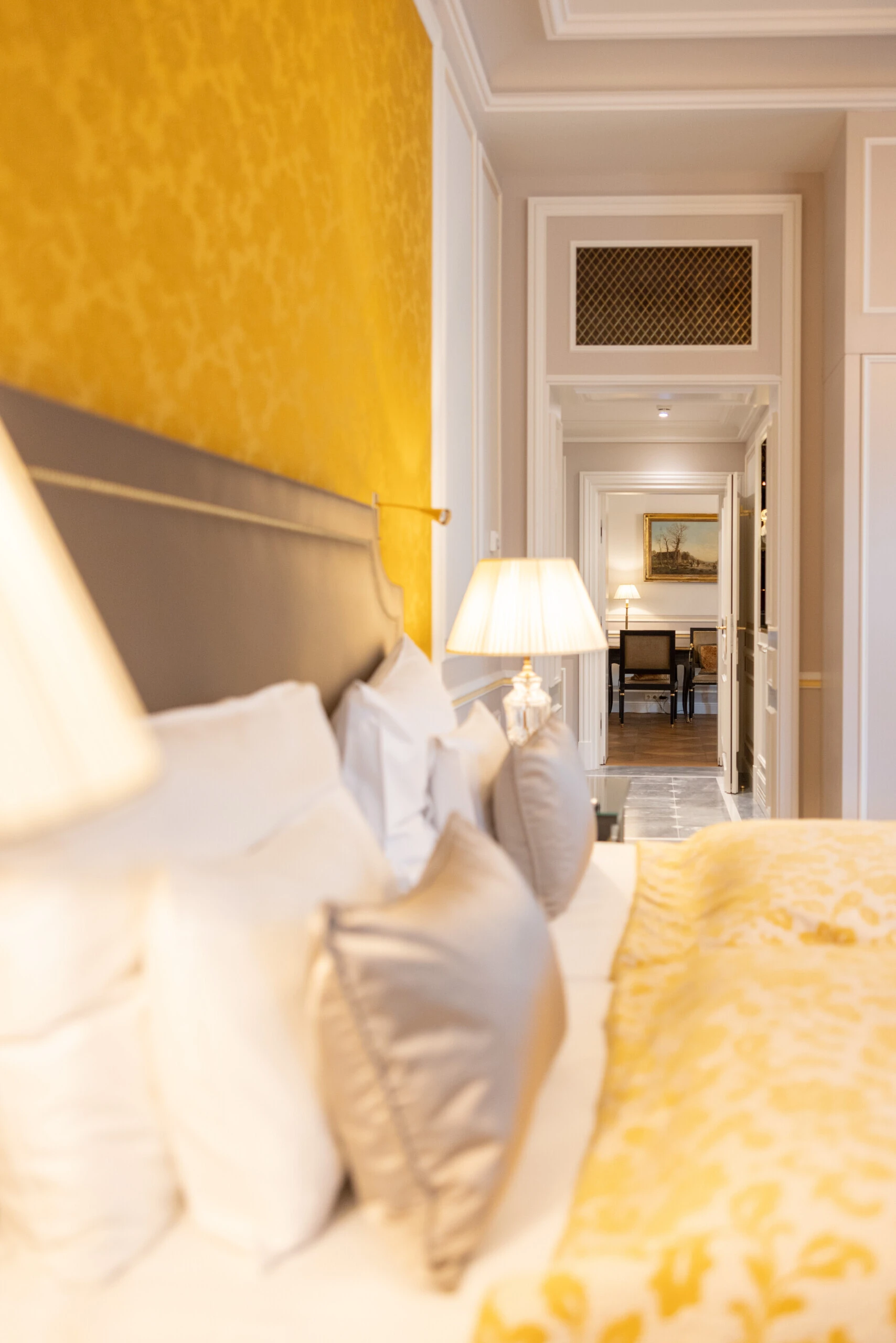 Blick vom Bett mit goldgelber Decke durch einen hellen Flur auf einen Schreibtischbereich, flankiert von klassischer Wandgestaltung und stilvoller Beleuchtung.
