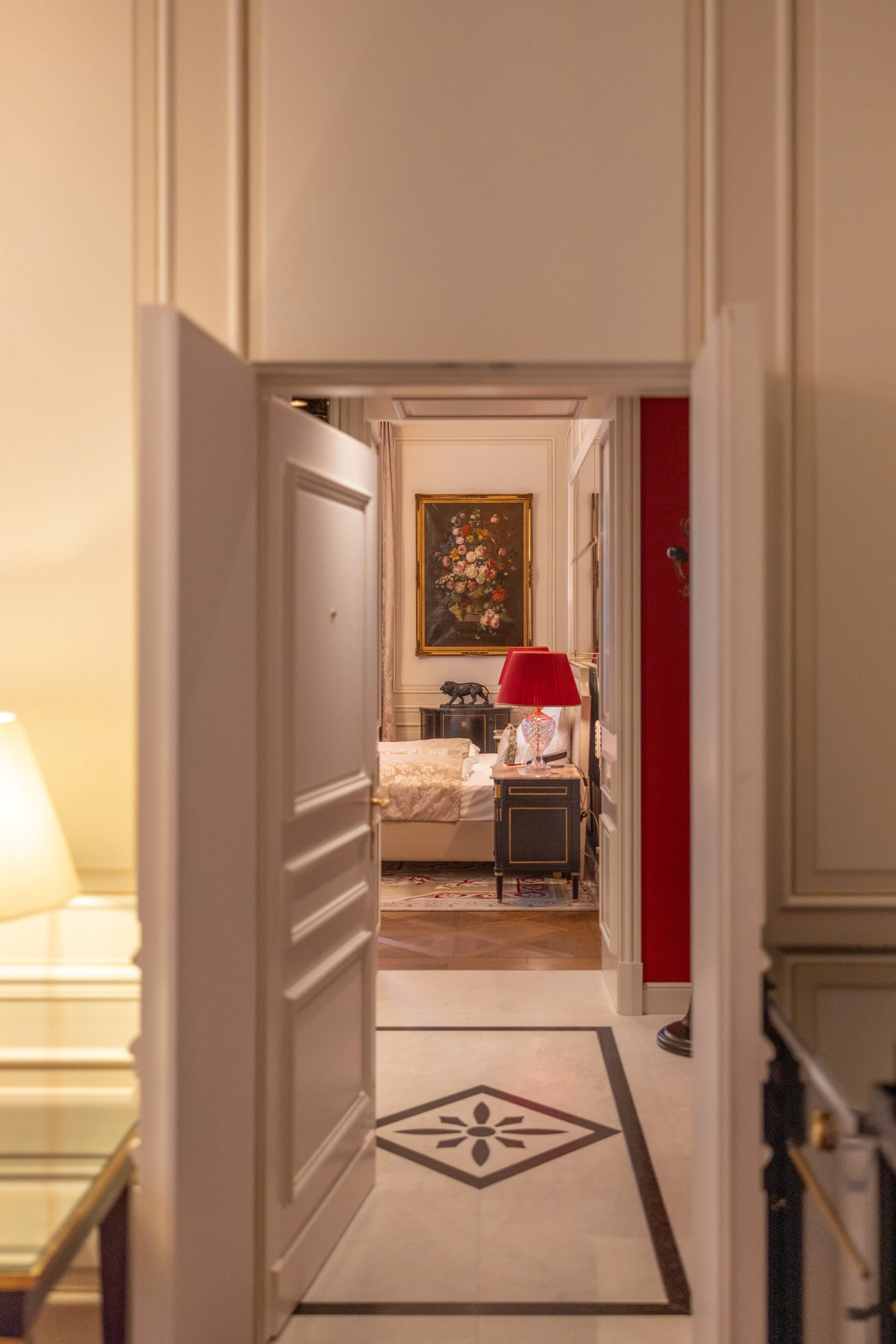 Durchblick auf ein stilvolles Schlafzimmer mit floraler Wandkunst, roter Tischlampe und klassischem Interieur, eingerahmt von hellen Wänden und Marmorboden mit Ornament.