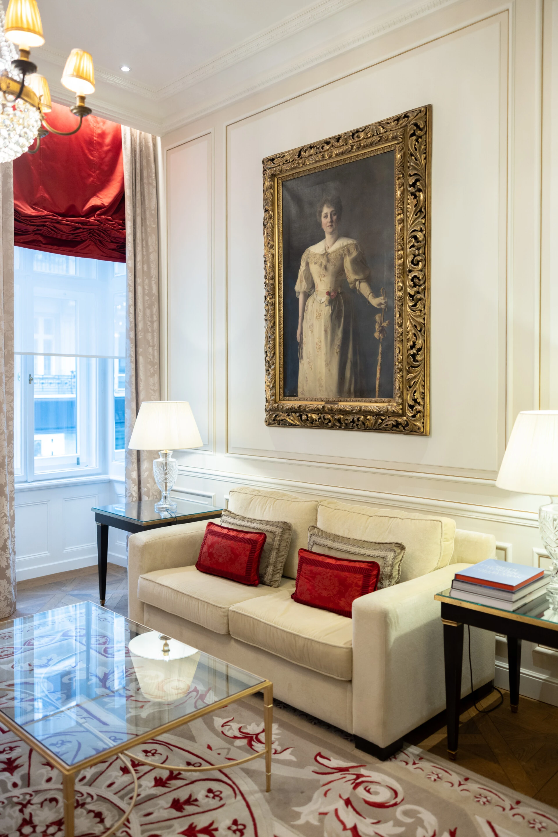 Eleganter Wohnbereich mit klassischer Couch, roten Kissen, Glastisch und großem Porträt in reich verziertem Rahmen an cremefarbener Wand.