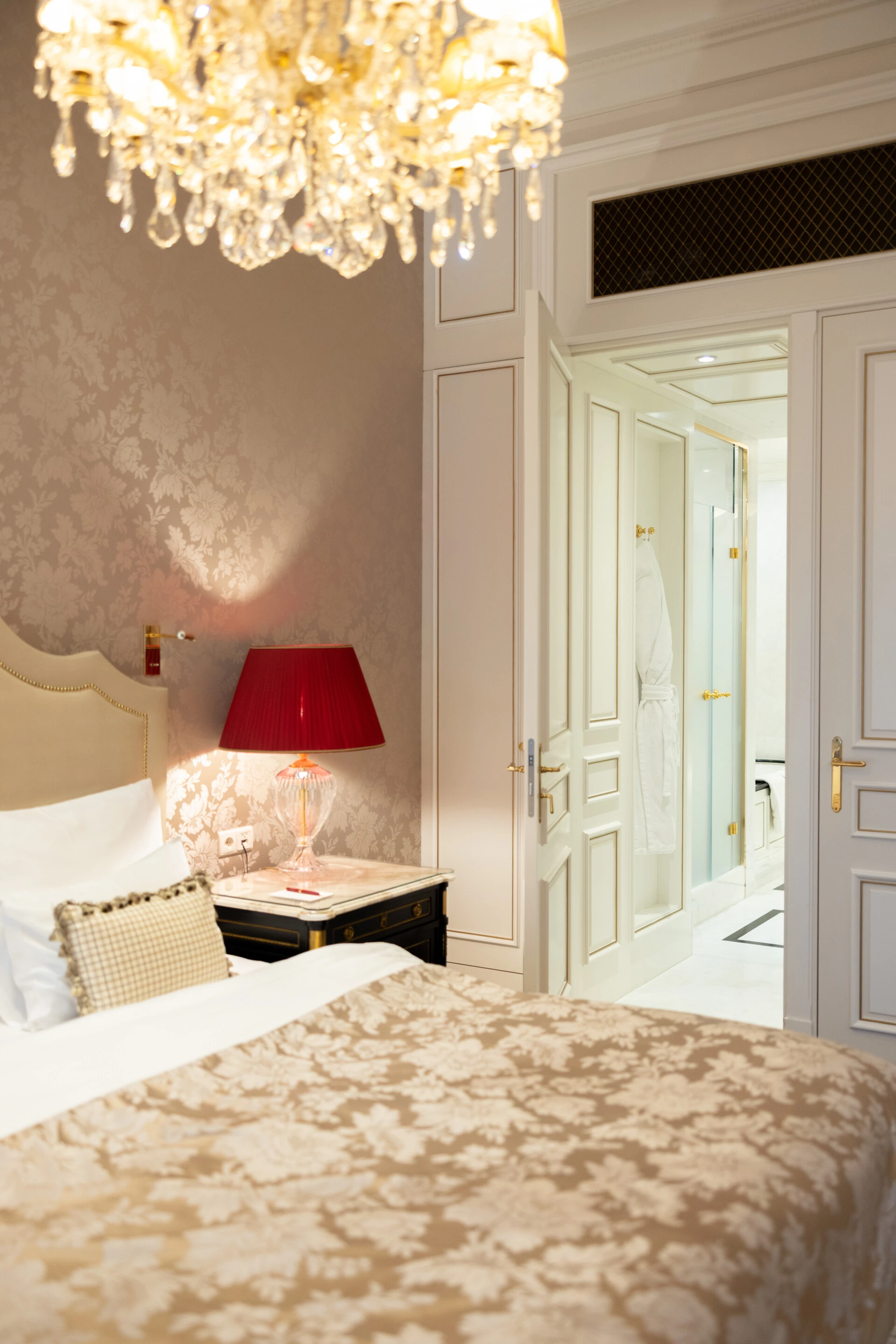 Teilansicht eines Schlafzimmers mit floraler Tapete, roter Tischlampe und Blick ins helle Badezimmer mit offenem Bademantel.