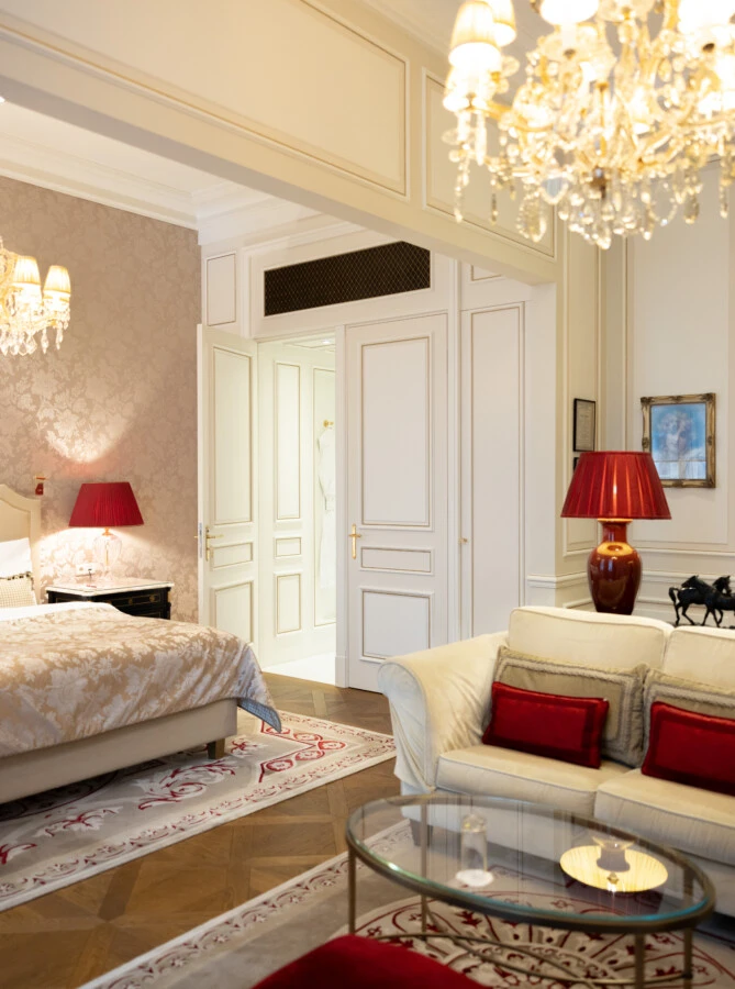 Luxuriöses Schlafzimmer mit cremefarbenem Bett, Kristalllüstern, roten Akzenten und eleganter Sitzgruppe auf floral gemustertem Teppich.