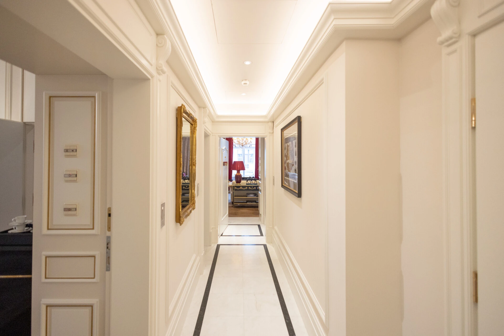 Heller Flur mit Marmorfliesen, dezenten Wandprofilen, goldgerahmten Bildern und Blick auf ein elegant möbliertes Zimmer mit roter Lampe und Kronleuchter.