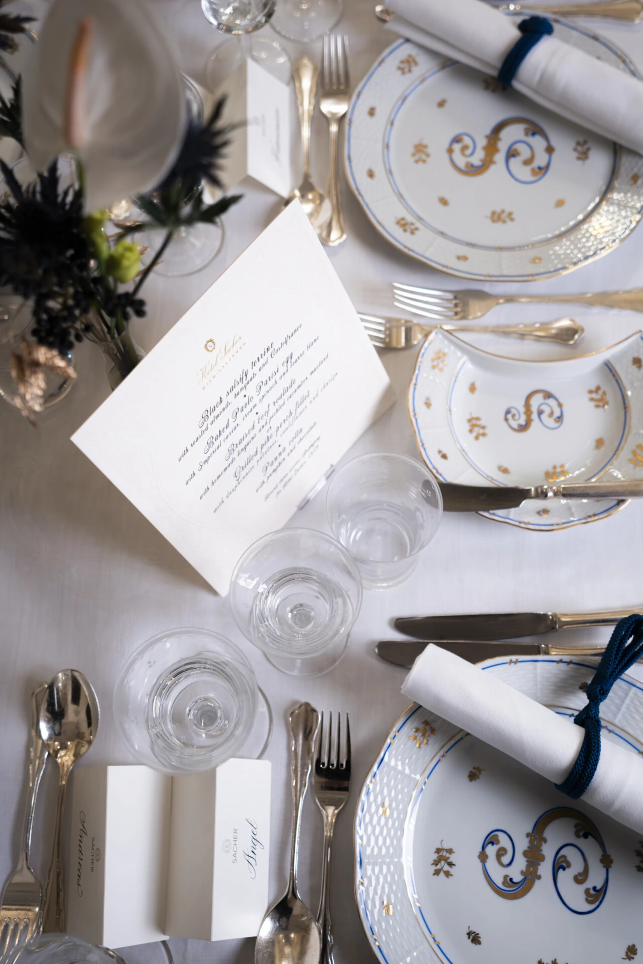 Elegant gedeckter Tisch mit weiß-goldenem Porzellan, blauer Serviettenbindung, Kristallgläsern und floraler Mitteldekoration.