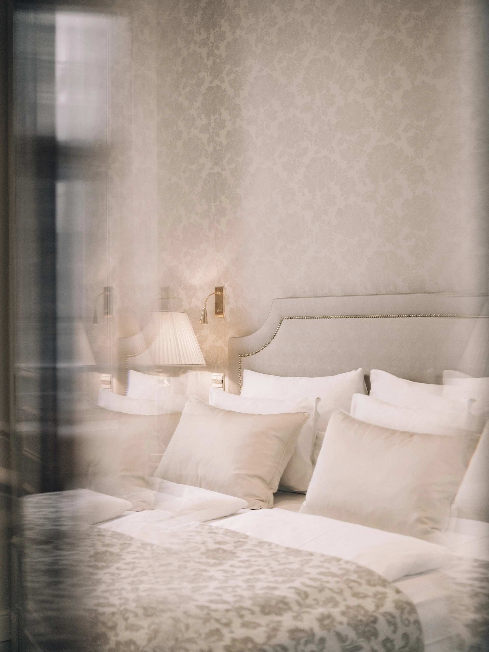 Ein elegantes Bett mit hellen Kissen und gemusterter Tagesdecke steht vor einer dezent verzierten Tapetenwand. Daneben befindet sich eine stilvolle Lampe auf einem Nachttisch.