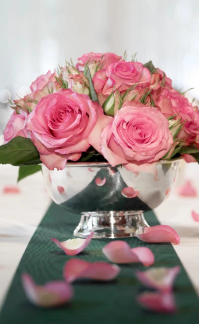 Tischdekoration mit rosa Rosen in einer silbernen Schale und verstreuten Blütenblättern auf einem grünen Tischläufer