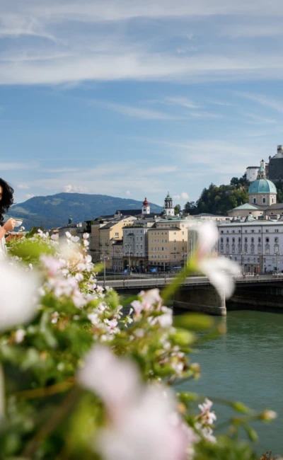 Frau mit Kaffeetasse auf Hotelbalkon mit Blumen im Vordergrund und Panoramablick auf Salzburg und die Festung Hohensalzburg