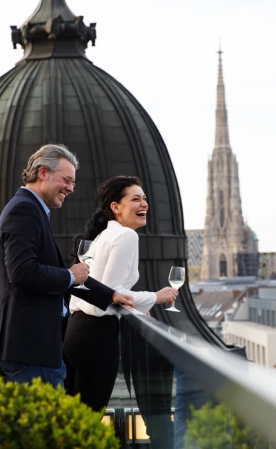 Ein Mann und eine Frau lächeln und lachen, während sie Weingläser in der Hand halten und die Aussicht von der Dachterrasse mit dem berühmten Stephansdom im Hintergrund genießen.