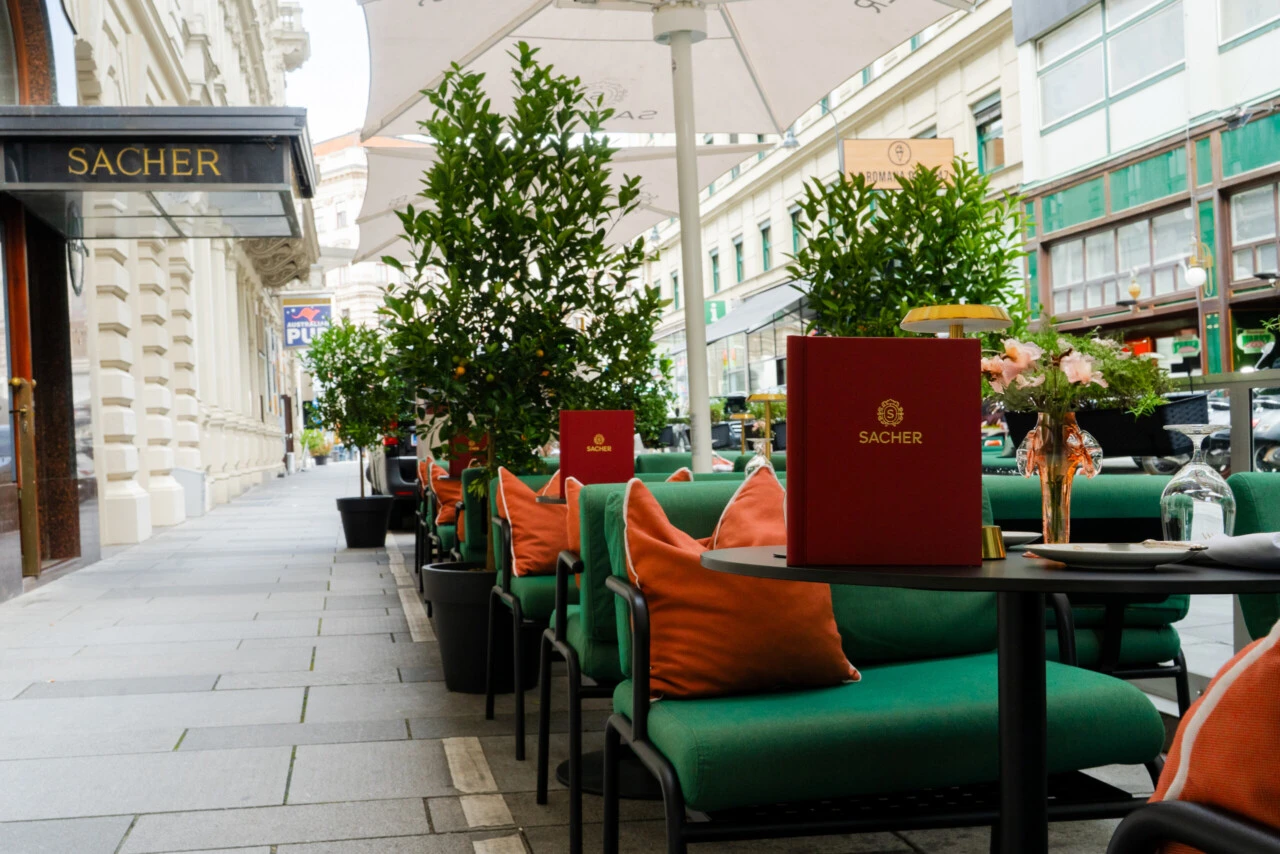Elegante Außensitzplätze im Sacher Summer Garden in Wien mit grünen Lounge-Stühlen, orangen Kissen und Zitronenbäumen entlang der Straße.