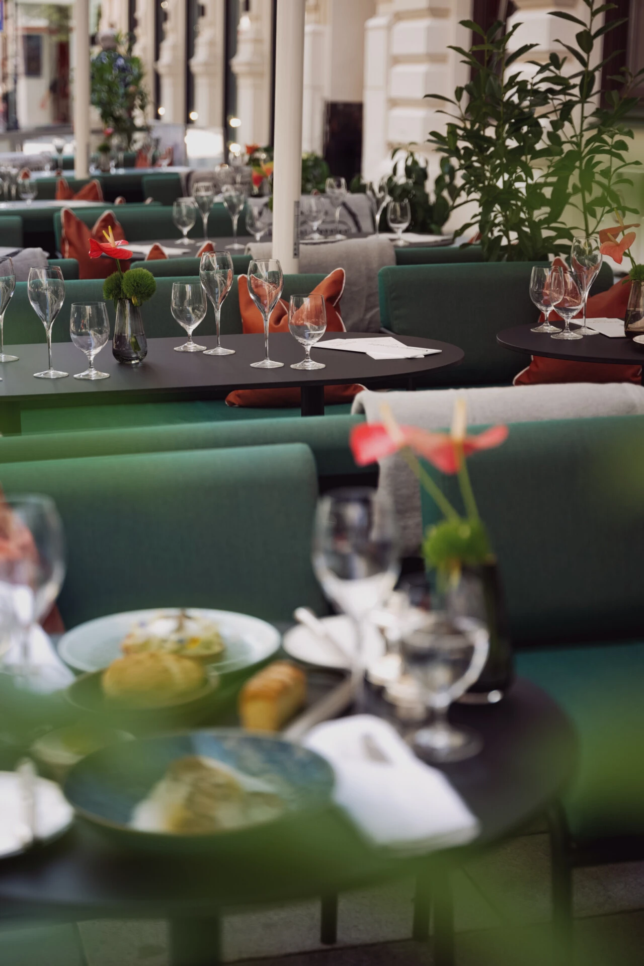 Elegante Tischdekorationen im Sacher Summer Garden mit Weingläsern, grüner und korallfarbener Bestuhlung, frischen Blumen und servierten Gourmetgerichten in entspannter Lounge-Atmosphäre.