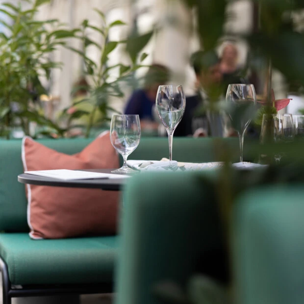 Nahaufnahme eines stilvoll gedeckten Tisches im Sacher Summer Garden mit Weingläsern, korallfarbenen Kissen und üppigem Grün für ein intimes, gehobenes Outdoor-Erlebnis.