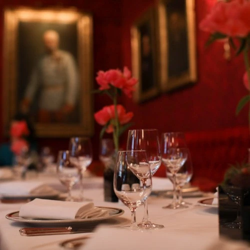 Elegant eingedeckte Tische mit weißen Tischdecken, Glasgarnituren und gefalteten Stoffservietten in einem stilvollen, rot tapezierten Raum mit goldgerahmten Porträts und rosa Pfingstrosen als florale Dekoration.