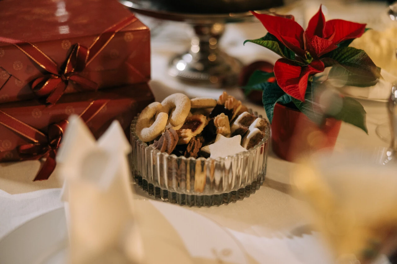 Weihnachtsplätzchen in Glasschale auf weißem Tischtuch. Im unscharfen Hintergrund Geschenke, Kuchen und Poinsettie.