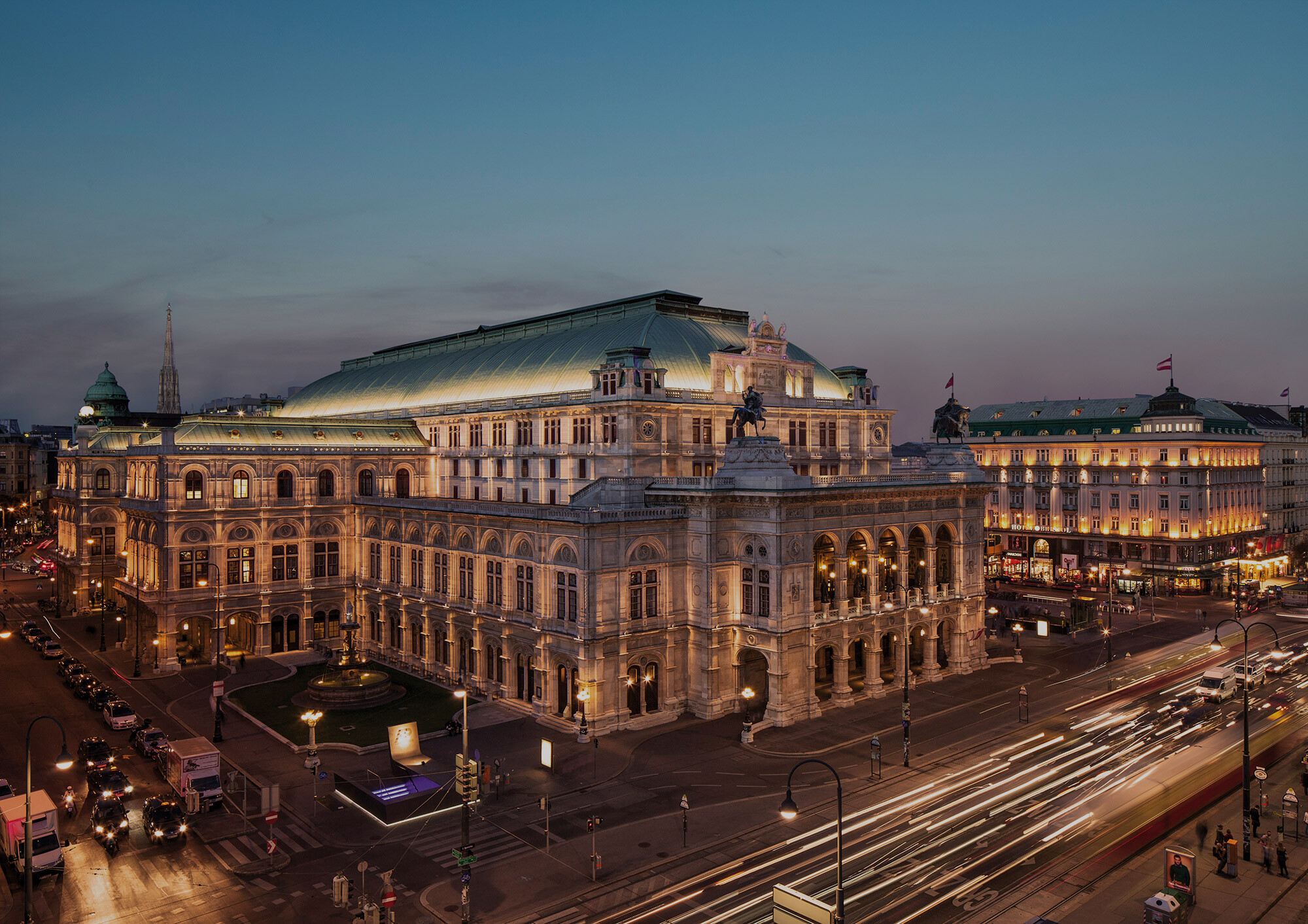 Die Wiener Staatsoper im Zentrum des Bildes ist prachtvoll beleuchtet, ihre neoklassizistische Fassade wirkt imposant und majestätisch. Rechts daneben das ebenfalls angestrahlte Hotel Sacher