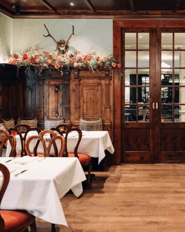 Rustikaler Speiseraum mit holzvertäfelten Wänden, festlich gedeckten Tischen und klassischen Stühlen. Über den Holzverkleidungen florale Gestecke und Hirschgeweihe. In der Mitte eine zweiflügelige Glastür. Der Raum trägt den Namen Zirbelzimmer“.