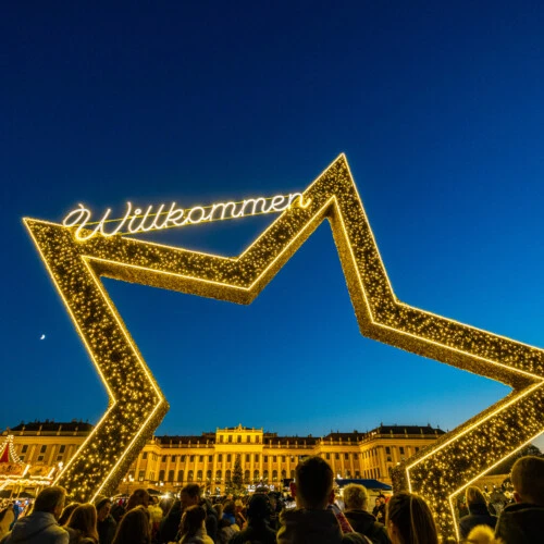 Ein riesiger beleuchteter Stern mit dem Wort „Willkommen“ steht vor einem großen, beleuchteten Gebäude während eines abendlichen Weihnachtsmarktes.