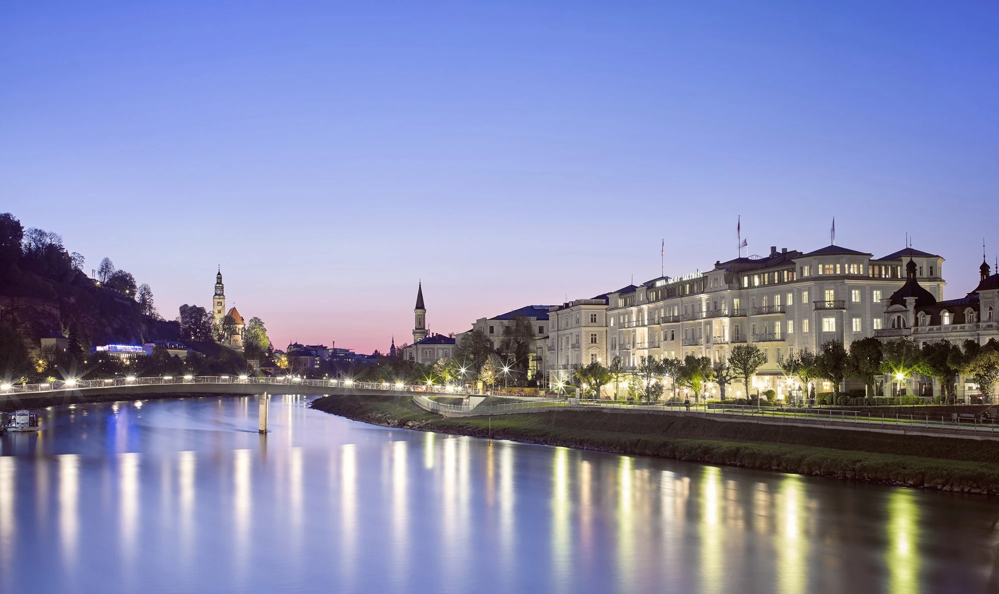 Abendliche Panoramaaufnahme der Salzburger Altstadt mit dem Hotel Sacher Salzburg im rechten Bildbereich, prachtvoll beleuchtet und spiegelnd in der Salzach.