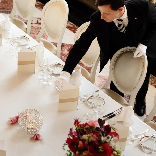 Ein eleganter gedeckter Tisch mit weißen Polsterstühlen, kunstvoll gefalteten Stoffservietten, Menü- oder Platzkarten und gläsernen Windlichtern. Ein Kellner in schwarzer Uniform mit weißen Handschuhen richtet präzise das Gedeck aus