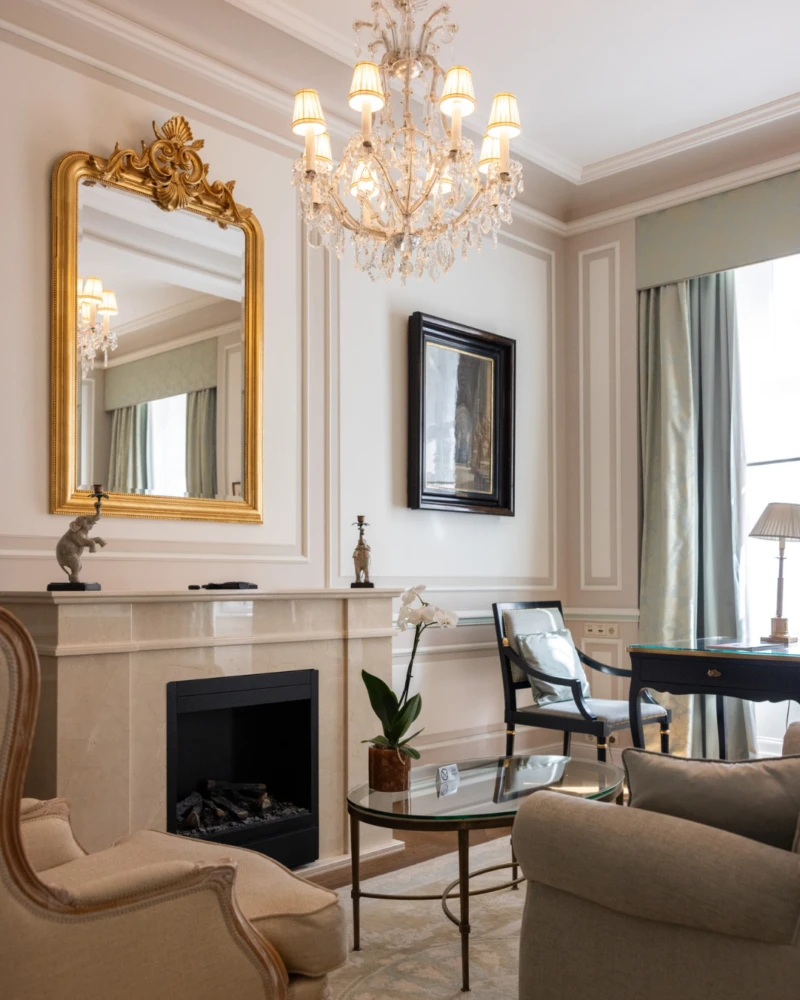 Ein klassisches Wohnzimmer mit Kamin, verziertem Goldspiegel, Kristallleuchter und eleganten antiken Möbeln.