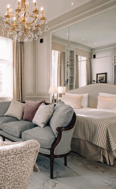 Eine luxuriöse Hotelsuite mit einem grauen Sofa, einem großen Bett und einer Spiegelwand, die das elegante Interieur widerspiegelt.