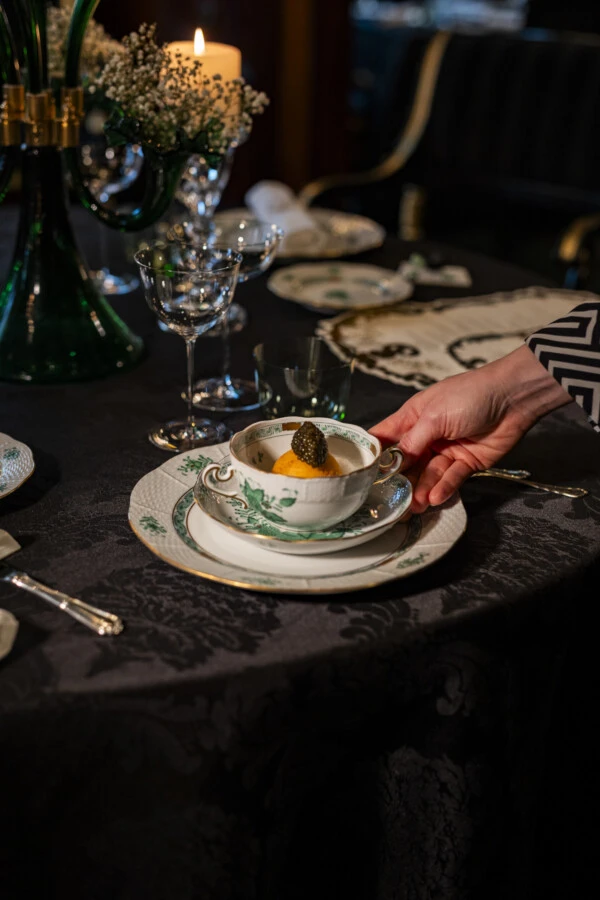Eine Hand serviert eine Schüssel Suppe mit einem orangefarbenen Inhalt und Kaviar, auf einem elegant gedeckten Tisch.