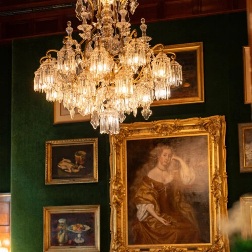 Ein goldener Kristallleuchter hängt vor einer grünen Wand, die mit verschiedenen gerahmten Gemälden, einschließlich eines großen Porträts einer sitzenden Frau, geschmückt ist.
