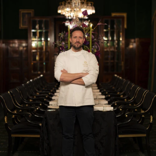 Ein Küchenchef in weißer Kochjacke und schwarzer Hose steht mit verschränkten Armen vor einem langen, elegant gedeckten Tisch unter einem Kronleuchter.