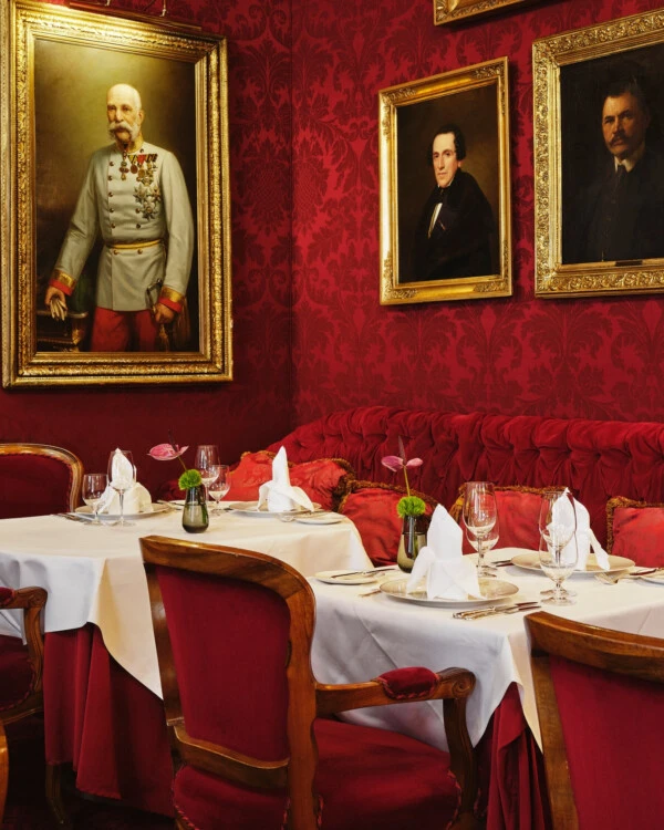 Elegantes Restaurant im Hotel Sacher Wien mit roten Samtstühlen, weißer Tischdekoration, Blumenschmuck und historischen Porträts an den Wänden.
