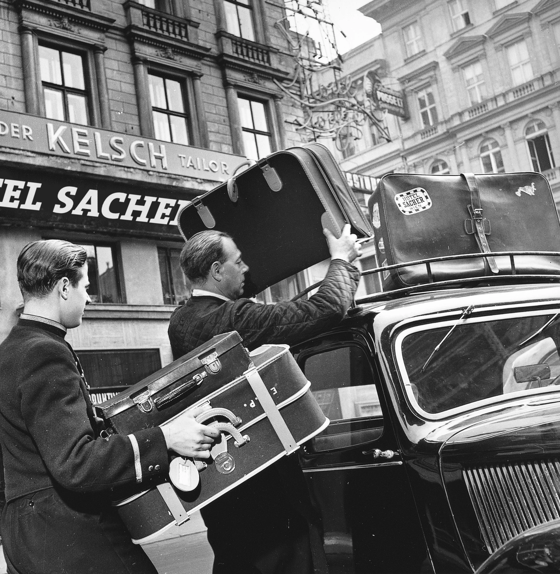 Schwarzweißfoto aus der Mitte des 20. Jahrhunderts: Zwei Männer – ein Hotelangestellter in Uniform und ein Gast – verladen stilvolle Lederkoffer auf das Dach eines Autos vor dem Hotel Sacher.