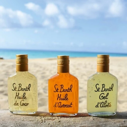 Drei Pflegeprodukte von Ligne St Barth – Kokosöl, Avocadoöl und Aloe Gel – auf einem Strand-Holzbalken mit Sand, Meer und blauem Himmel im Hintergrund.