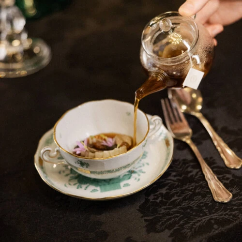 Hand gießt klare Brühe aus Teekanne in eine Schale mit Einlage und Blüten.