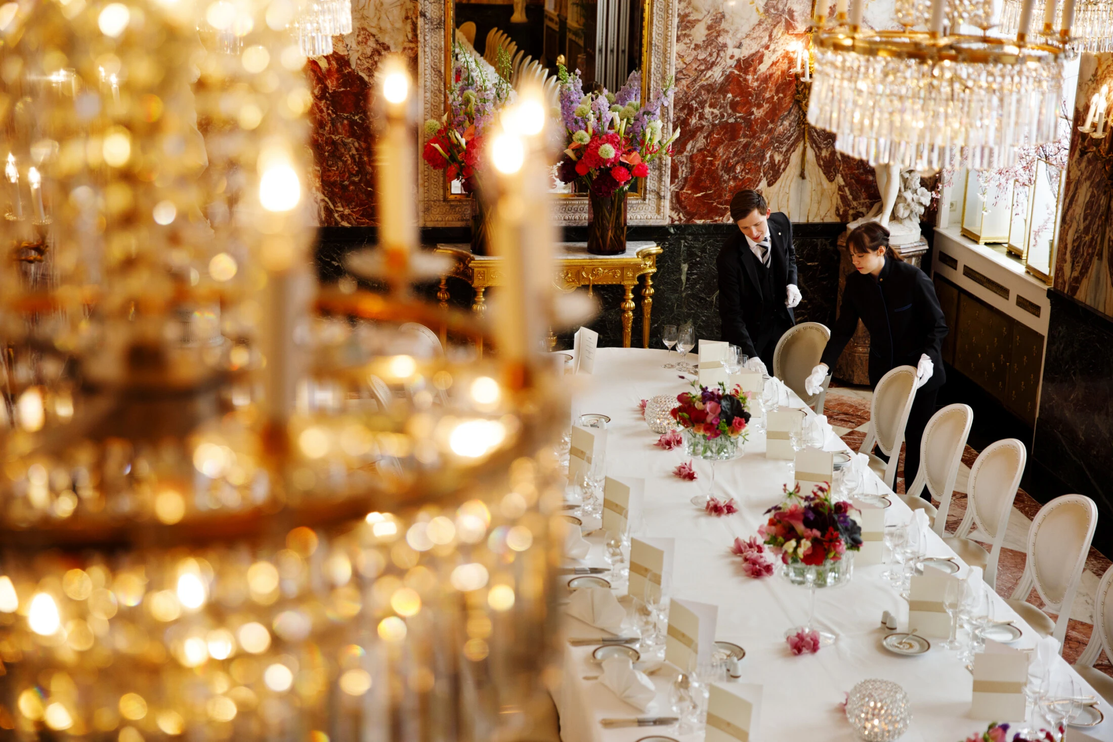 Unter glitzernden Kronleuchtern und prächtigen Marmorwänden bereiten zwei Mitarbeiter in formeller Kleidung im Hotel Sacher Wien sorgfältig einen opulenten Esstisch vor, der mit frischen Blumen und eleganten Gedecken geschmückt ist.