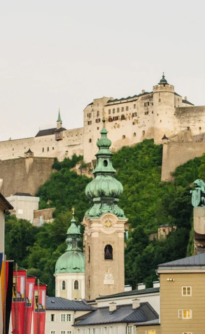 Die Festung Hohensalzburg dominiert die Skyline, gesehen von einer Salzburger Straße mit Kirchtürmen und bunten Flaggen.